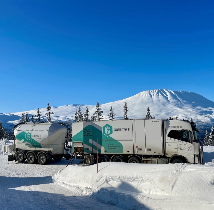 Gulvavrettingsbil og tilhenger står parkert i snødekt landskap med fjell i bakgrunnen.